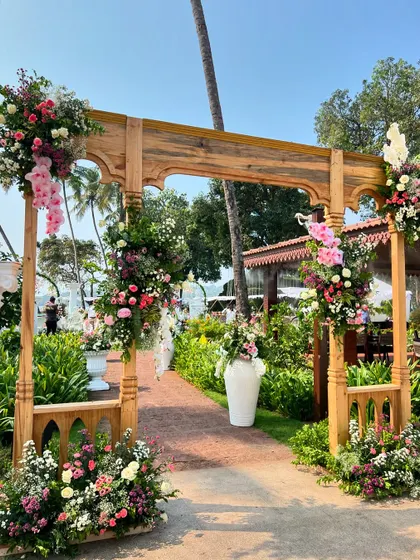 A handcrafted wooden gate adorned with lush pink and white florals, creating a beautiful entrance for a 'Roman Fiesta' themed mehendi by the beach in Goa.