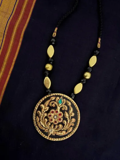 This elegant black and gold pendant showcases intricate floral gesso work, highlighted with real Tanjore gold foil and a single gemstone. It is paired with brushed gold and black beads for a sophisticated look.