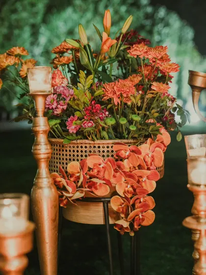A beautiful arrangement of orange orchids and chrysanthemums in a cane planter, surrounded by the warm glow of candles in tall, elegant holders. This adds a touch of warmth and nature to the glam setup.