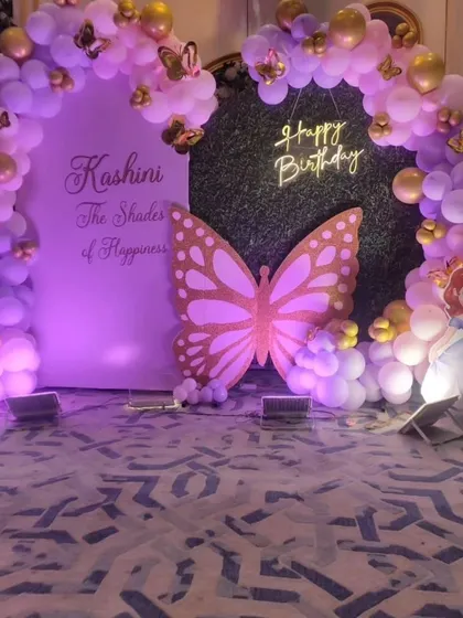 A close-up of a butterfly and princess-themed backdrop. This setup for Kashini's birthday combines a green foliage panel with a personalized board, framed by a purple and gold balloon arch and flanked by Sofia the First cutouts.