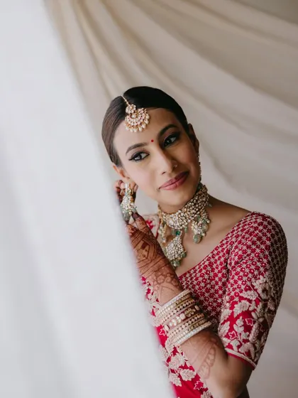 A beautiful close-up of a bride adjusting her earring, looking at the camera with a gentle smile, framed by a soft white curtain.