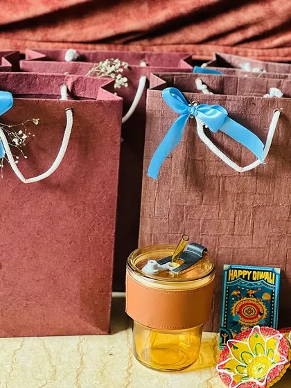 A few of the brown paper gift bags displayed with some of the items that go inside, like a glass tumbler and decorative diyas.