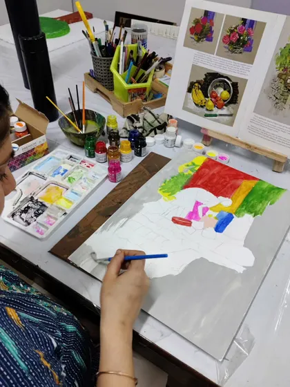 A student works on her gouache painting, blocking in areas of color while referencing an art book.