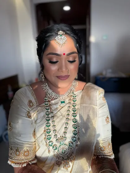 A moment of calm before the ceremony. This bride's Muhurtham look features soft, smoky eyes and a neutral lip, beautifully complementing her off-white and gold saree and stunning emerald jewelry.