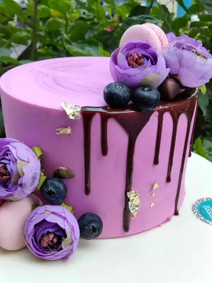 A minimal yet striking purple cake with a dark chocolate drip. The cake is decorated with fresh blueberries, macarons, and purple flowers for a rich and elegant look.