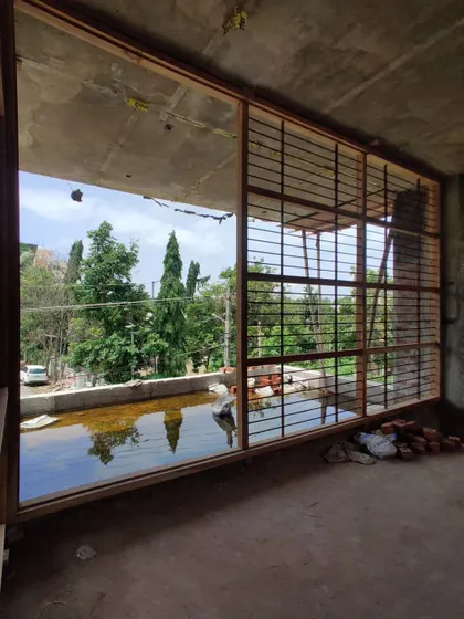 A large window frame installed on an upper floor, capturing reflections of the sky and trees in a pool of rainwater on the floor.