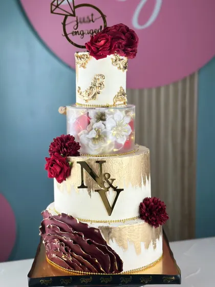 A touch of drama, a whole lot of elegance. This tall, four-tiered cake combines a rich maroon with brushed gold, featuring a clear tier filled with glowing lights and handcrafted flowers.