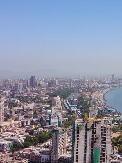 A daytime drone panorama showing the curve of Marine Drive in Mumbai, with the city on one side and the Arabian Sea on the other.