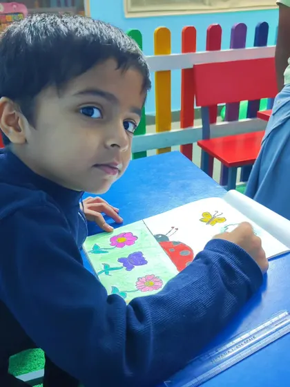 A student looks up from his work, coloring a ladybug drawing at his desk in our colorful classroom.