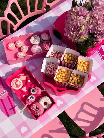 An overhead view of a kids' table, featuring pink gingham tablecloths and an array of themed treats like donuts, cupcakes, and popcorn.