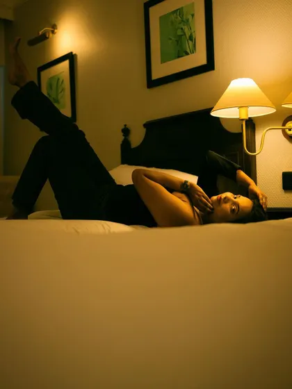 A playful and stylish portrait of a woman lying on a hotel bed, illuminated by warm lamplight. The composition and lighting create a relaxed yet glamorous feel, perfect for an indoor fashion shoot.