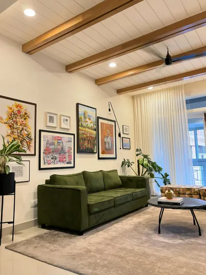A full view of the living room with its vaulted ceiling, wooden beams, and extensive gallery wall. The neutral rug grounds the space, allowing the green sofa and colorful artwork to stand out.