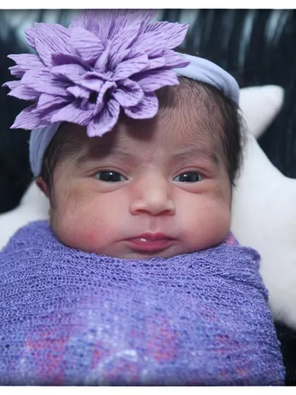 A beautiful close-up of a newborn baby girl with a lovely purple flower headband, wrapped in a matching purple swaddle.