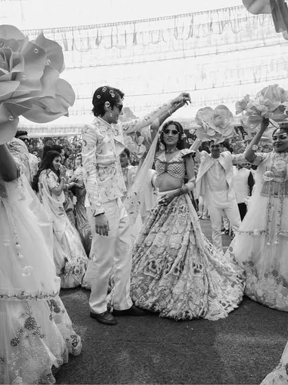 The bride's grand entrance at her carnival-themed event, surrounded by dancers with giant paper flowers. This black and white shot emphasizes the drama and excitement of the moment.