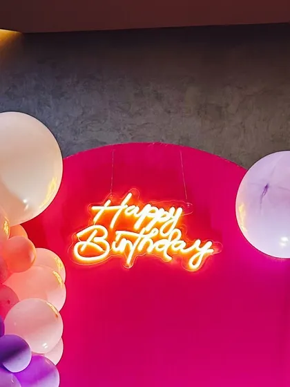 A bright orange "Happy Birthday" neon sign hanging against a bold magenta backdrop, surrounded by balloons.