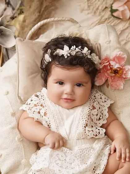 A beautiful baby girl in a white lace romper and floral headband smiles sweetly while lying in a basket, surrounded by soft fabrics and flowers.