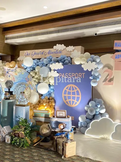 A full view of the travel-themed stage, with a large passport backdrop, balloon clouds, and props like a Vespa and vintage luggage.