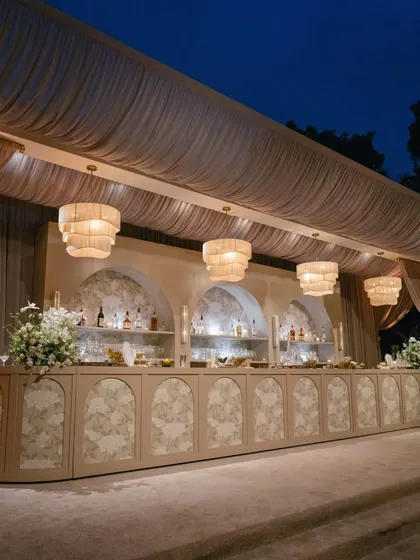 A stunning, custom-designed bar for an all-white party. The long bar features floral-patterned panels and arched back-shelves, illuminated by a row of contemporary chandeliers.