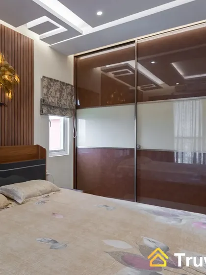 A master bedroom featuring a sliding wardrobe with brown lacquered glass doors. The accent wall behind the bed has fluted wood paneling and a decorative golden tree of life.