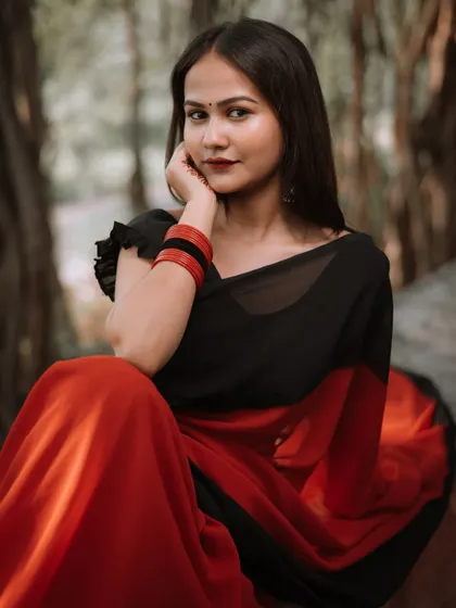A classic seated pose in a beautiful red and black saree. The natural forest-like background adds a rustic feel to this ethnic portrait, highlighting the simple elegance of the attire.