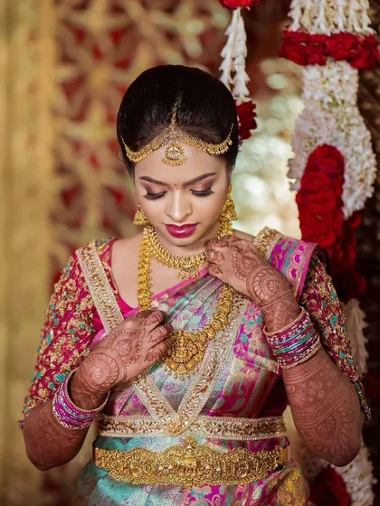 The bride adjusting her necklace, a moment that shows the complete look with her traditional gold jewelry. The makeup is designed to enhance, not overpower, these beautiful elements.