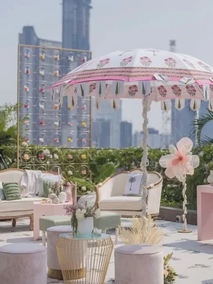 A chic and modern rooftop Mehendi in an urban setting. The pastel pink and white decor, elegant seating, and custom-printed umbrella create a sophisticated and stylish ambiance with a stunning city skyline view.