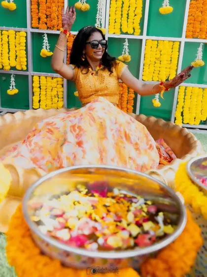 A playful moment from a Haldi ceremony. The bride, seated in a decorative basin, joyfully tosses flower petals, surrounded by the bright colors of the event.