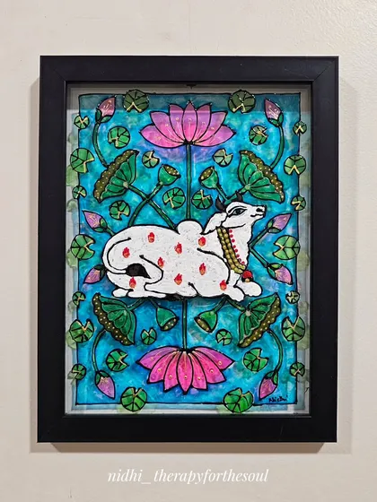 This framed Pichwai-style cow, painted on glass, is surrounded by lotuses. The glass medium gives the colors a special luminosity, making it a vibrant addition to any wall.
