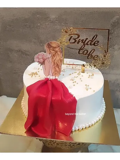 A chic and stylish "Bride to be" cake. The design includes a beautiful illustration of the bride, a flowing red fondant dress, and a custom gold topper, all on a base of elegant white buttercream.