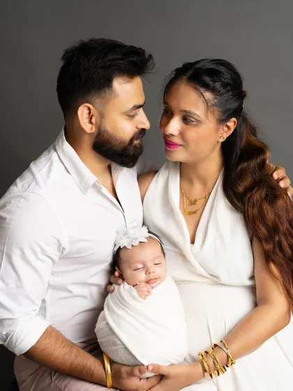 A beautiful color portrait of a new family. The parents look at each other with love while holding their peacefully sleeping newborn, captured against a simple grey studio background.