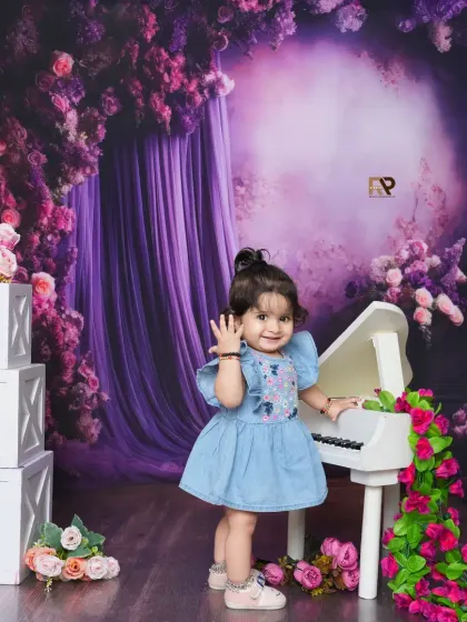 A smiling baby girl waves from her spot at the mini piano in a beautiful floral setup. The "ONE" blocks indicate a special first birthday celebration.