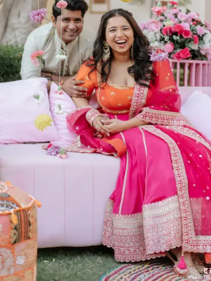 The bride shares a laugh with the groom during their colorful Mehendi ceremony, seated on a beautifully decorated pink sofa.