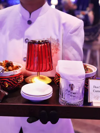 A server presents a tray of "Five Spice Baby Corn & Broccoli," served in ornate bowls with a decorative figurine. This theatrical presentation transforms a simple dish into a memorable experience.