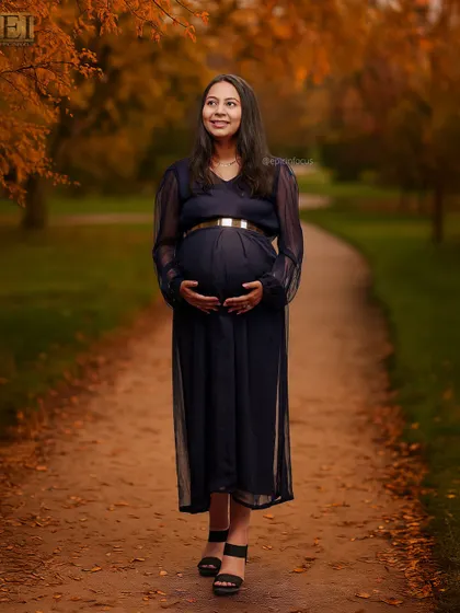 A full-length shot of a mom-to-be in a sophisticated navy blue gown, walking along a path in an autumn-themed outdoor setting.