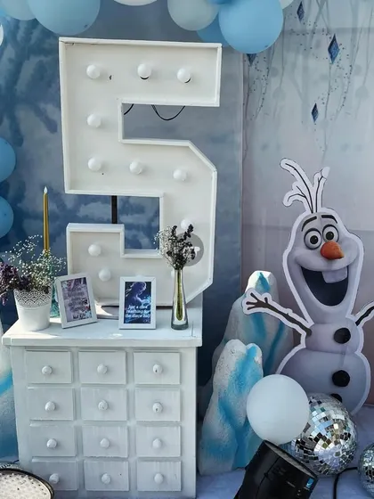 A friendly Olaf cutout stands next to a lit-up number '5' and a white dresser, creating a charming corner at the Frozen-themed celebration.