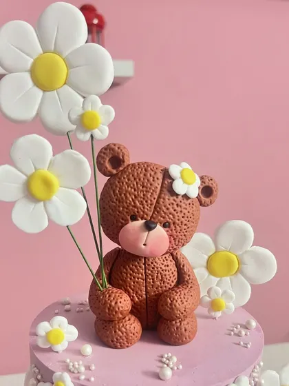 A close-up of the handcrafted teddy bear and daisy topper. I love adding texture and small details, like the flower in the bear's hair, to make each figurine special.