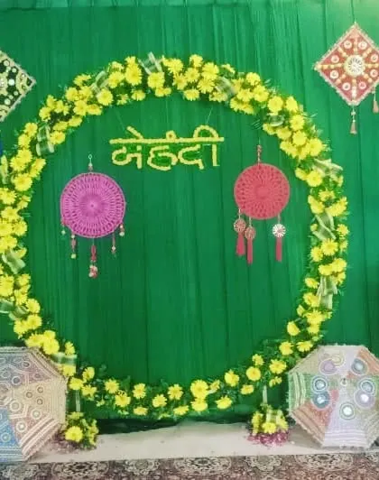 A simple Mehendi photo booth with a circular frame of yellow flowers against a green backdrop. The decor includes hanging dreamcatchers and colorful umbrellas.