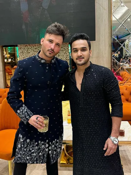 Two friends in complementary black and navy blue ethnic outfits. One wears a heavily embroidered bandhgala, while the other is in a simpler, textured black kurta.