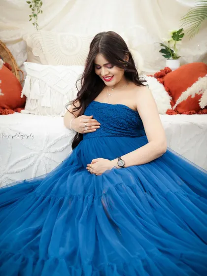 A serene portrait of a mom-to-be in a deep blue tulle gown. She is seated in a cozy boho setting, looking down at her bump with love.