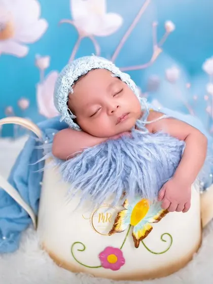 A sleeping newborn in a blue bonnet, nestled inside a decorative watering can prop against a dreamy blue floral background.