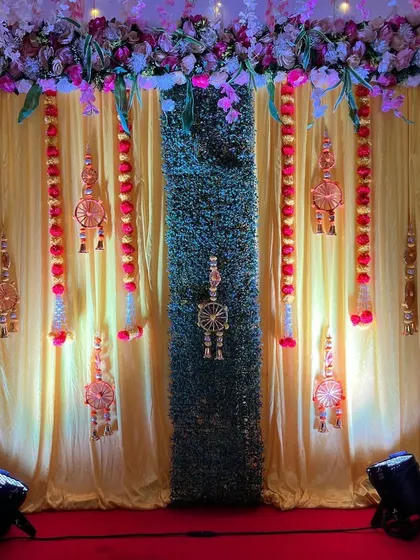 A classic Haldi backdrop with rich yellow curtains and a central artificial grass panel. The setup is decorated with traditional hangings and marigold flower strings, illuminated by spotlights for a warm glow.
