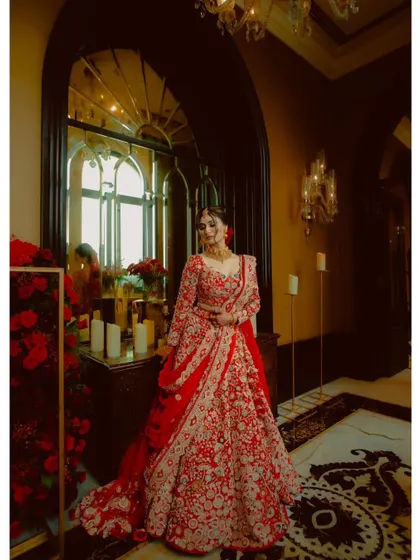 A bride looking absolutely regal in her red lehenga, surrounded by candles and flowers in a luxurious hotel setting.