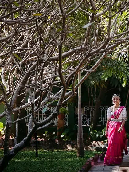 A bride in a bright pink saree walks through a grove of trees, a beautiful shot that highlights the natural beauty of the venue.