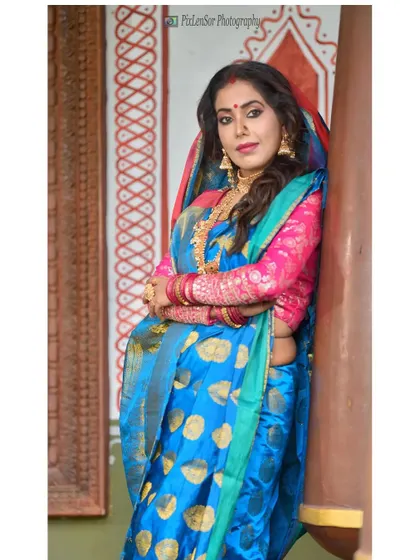 An actress styled in a Bengali look, leaning against a pillar. The gold jewellery shines against the vibrant blue and pink saree.