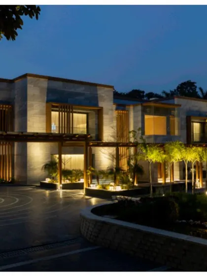 The farmhouse exterior illuminated at night. The landscape lighting design highlights the architectural features, the circular driveway pattern, and the surrounding trees, creating a serene and dramatic ambiance.