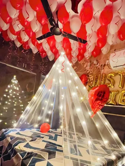 A cozy and romantic room decoration featuring a tent made of sheer fabric and fairy lights over the bed. The ceiling is filled with red and white balloons, and 'Just Married' foil letters are placed on the headboard.