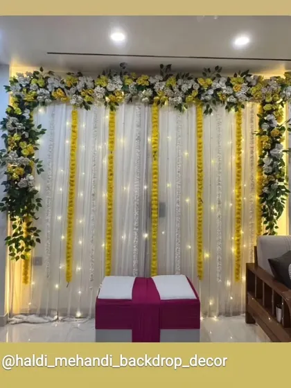 An elegant indoor Haldi backdrop with white and yellow floral arrangements. The setup features a white curtain background with hanging marigold strings and fairy lights, perfect for home-based functions.