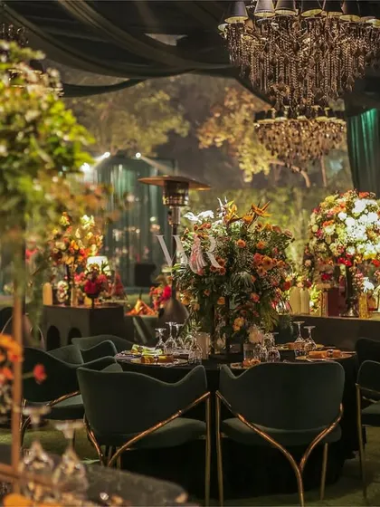 A view of the dining area, where tables are set with lush, colorful floral arrangements and surrounded by deep green velvet chairs. The layering of chandeliers and soft spotlights creates depth and a sense of luxurious intimacy.
