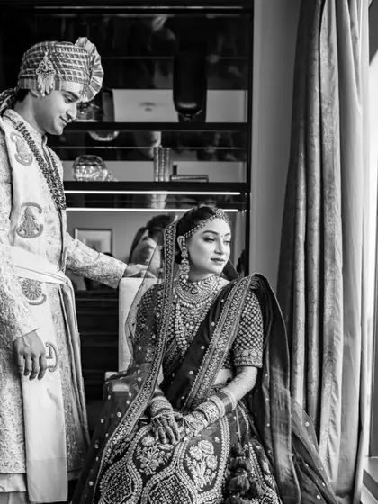 A timeless black and white portrait of the couple. The groom stands by his seated bride, creating a classic, regal composition by the window.