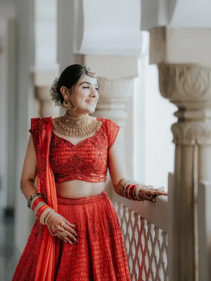 A radiant bride in a vibrant red lehenga, smiling as she looks out from a balcony. The natural light beautifully illuminates her features and her happiness.